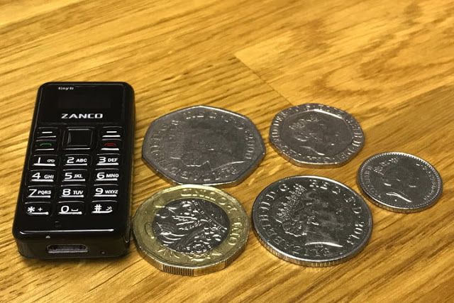 The image shows Zanco Tiny t1 placed on a table along with some coins to compare its size and give an idea of the tiny dimensions of the phone to the users